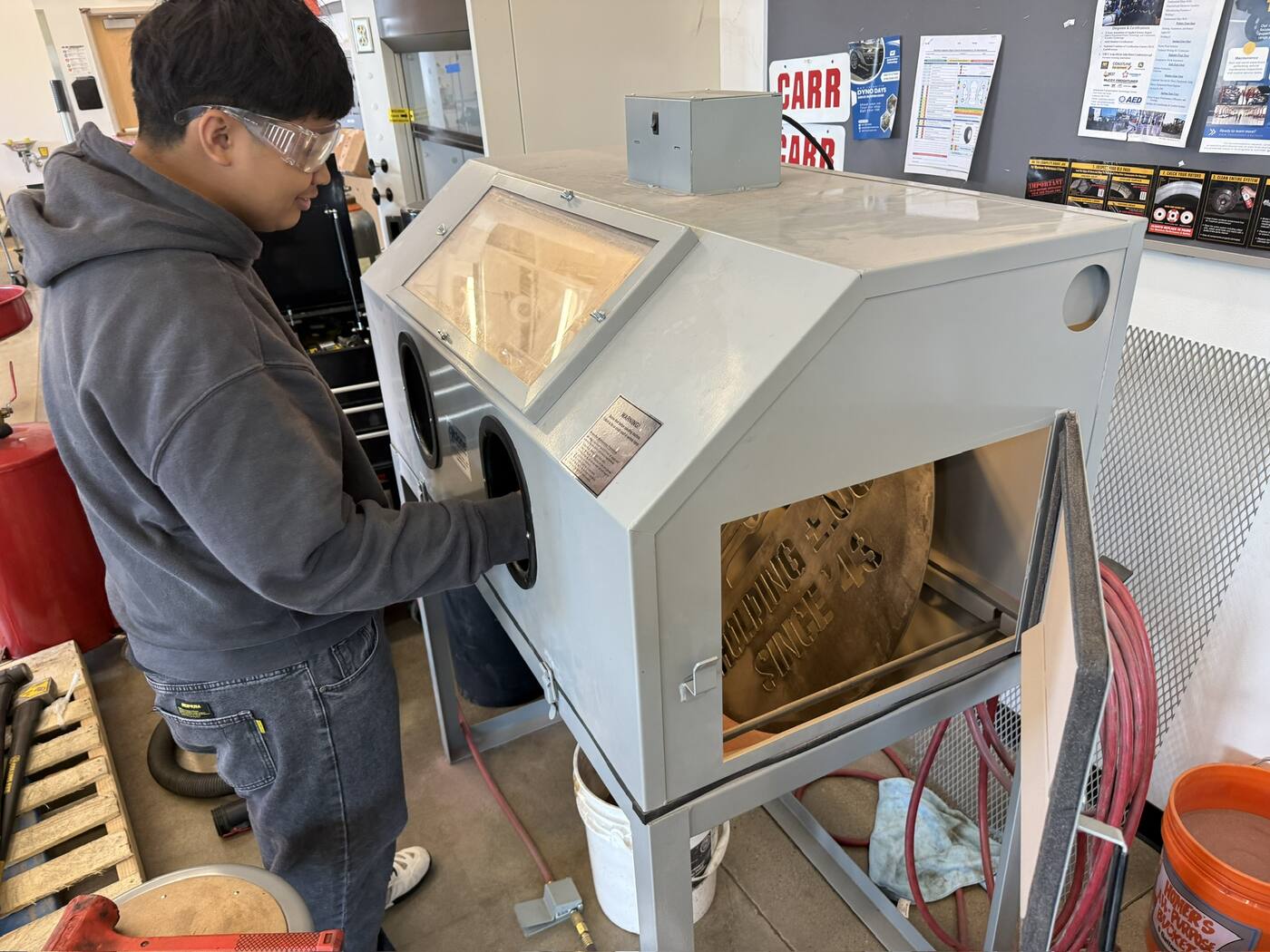 A student in safety glasses works at the grant-funded Cyclone blast cabinet. Through the open door, the steel sign sits inside, its surface already blasted to a clean, even finish.