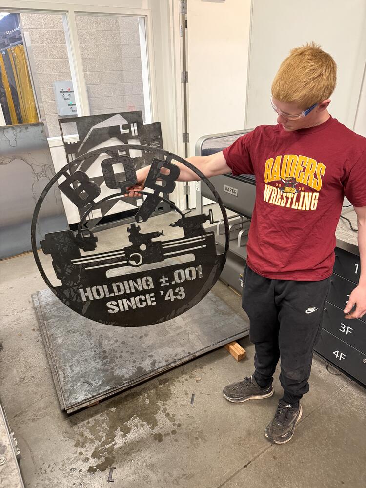 A student lifts the freshly plasma-cut steel sign off the cutting table. The metal is raw and dark with mill scale, and the lettering and lathe shapes are cut clean through.