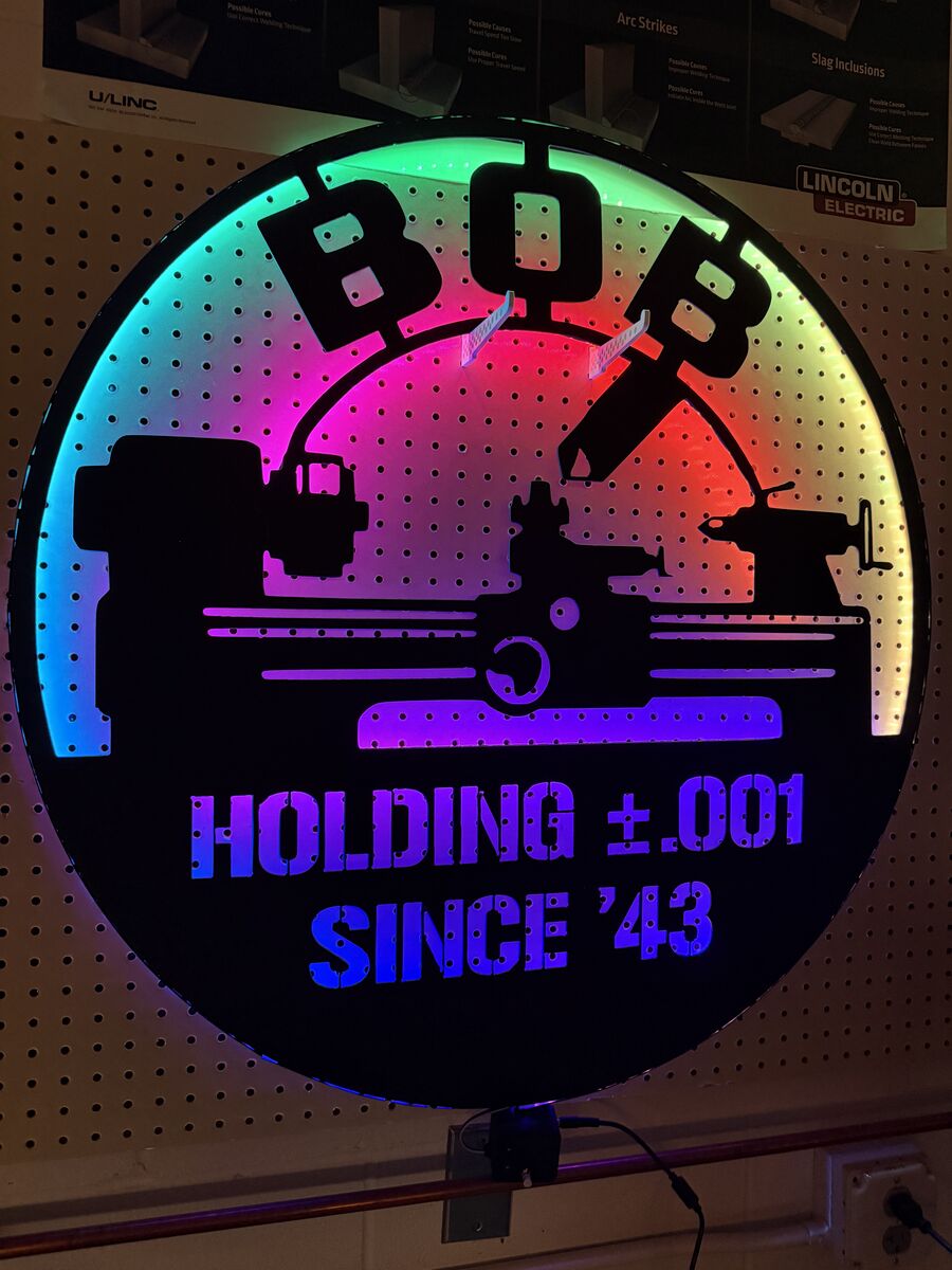 The finished sign lit up close: black powder-coated steel with BOB, a lathe, and HOLDING plus or minus .001 SINCE '43 standing out against bands of shifting LED color.