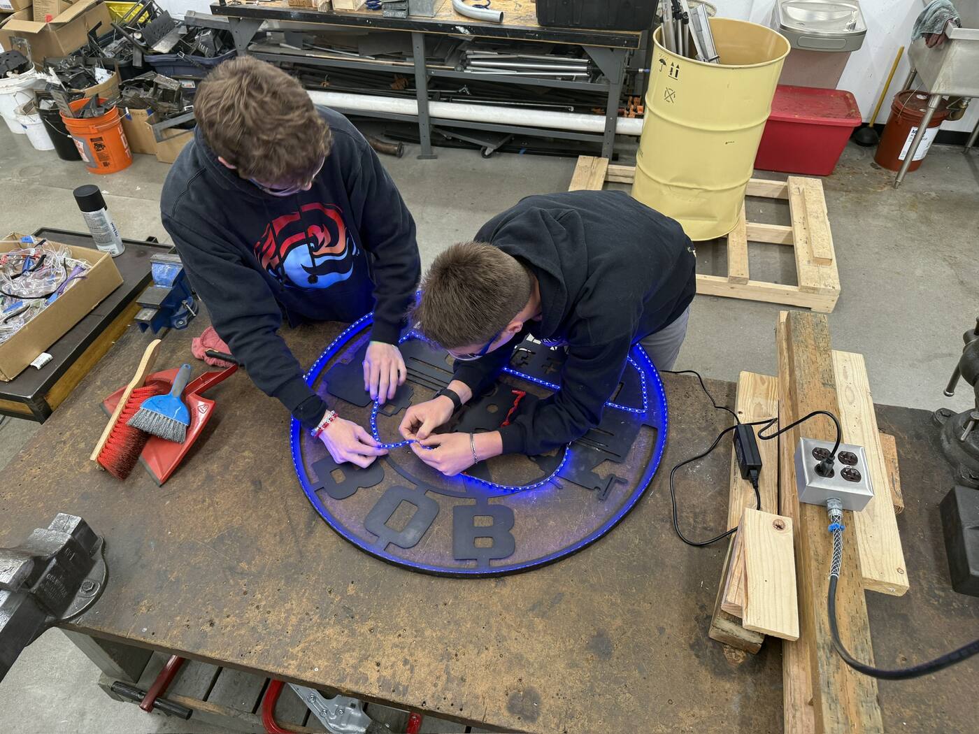 Two students lean over a workbench, fitting and soldering a blue LED strip around the inside rim of the powder-coated sign.