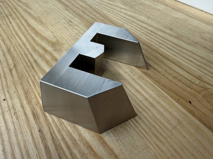 A small aluminum block machined with angled faces and a stepped notch, sitting on a wooden bench.